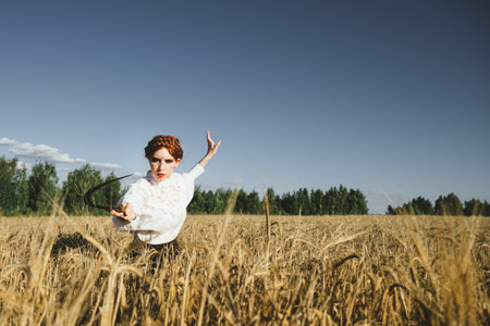 Slavic girl in a white dress and a black skirt with witch makeup stands in a field of rye and holds a terrible sickle in her hands. The concept of a mystical harvest.の写真素材