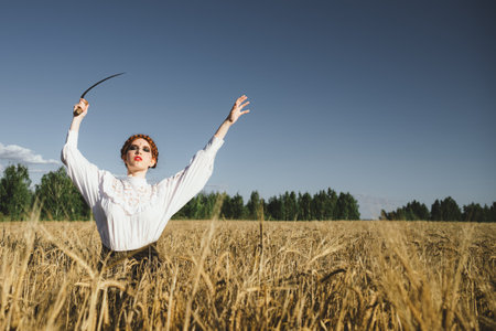 Slavic girl in a white dress and a black skirt with witch makeup stands in a field of rye and holds a terrible sickle in her hands. The concept of a mystical harvest.の写真素材
