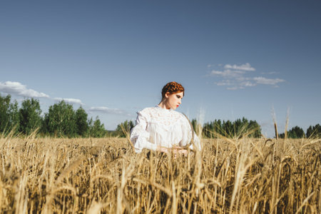 Slavic girl in a white dress and a black skirt with witch makeup stands in a field of rye and holds a terrible sickle in her hands. The concept of a mystical harvest.の写真素材