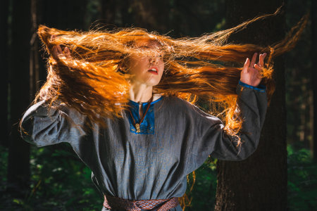 Red haired young woman in a linen blue dress is sitting in the middle of the forest in the rays of the sun. The girl emotions of despair left alone in the forest.の写真素材