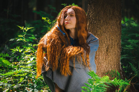 Red haired young woman in a linen blue dress is sitting in the middle of the forest in the rays of...