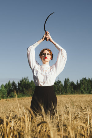 Slavic girl in a white dress and a black skirt with witch makeup stands in a field of rye and holds a terrible sickle in her hands. The concept of a mystical harvest.の写真素材