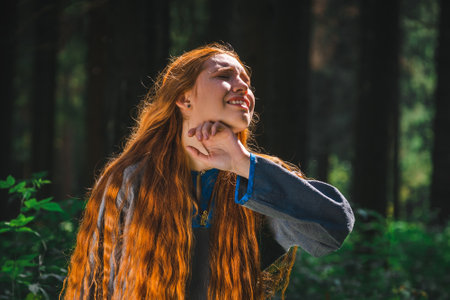Red haired young woman in a linen blue dress is sitting in the middle of the forest in the rays of the sun. The girl emotions of despair left alone in the forest.の写真素材