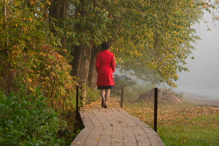 Red-haired woman in a red long raincoat stands on a wooden path in the countryside in autumn against...