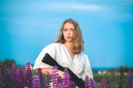 Happy woman in a white dress stands in a field with lupine flowers holding an automatic assault rifle.の写真素材