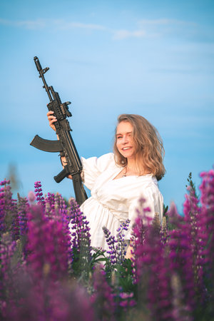 Happy woman in a white dress stands in a field with lupine flowers holding an automatic assault...