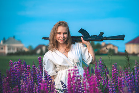 Happy woman in a white dress stands in a field with lupine flowers holding an automatic assault rifle.の写真素材