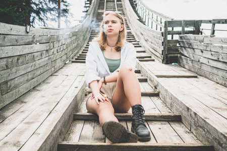 Young woman in casual clothes is sitting on an abandoned trampoline and smoking a cigaretteの写真素材
