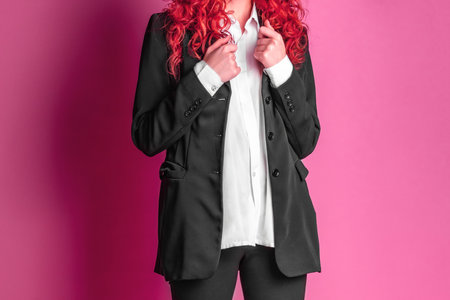 Studio photo of a young business woman in a black suit and white shirt on a pink background. A red haired businesswoman.の写真素材