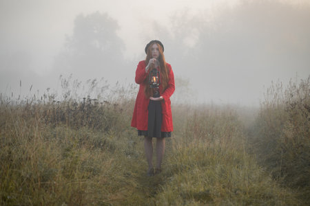 Woman with kerosene lamp in a red coat stands in a mystical misty morning field.の写真素材