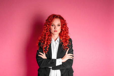 Studio photo of a young business woman in a black suit and white shirt on a pink background. A red haired businesswoman.の写真素材