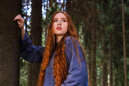 Red haired young woman in a linen blue dress is escape in the middle of the forest in the rays of the sun. The girl emotions of despair left alone in the forest.の写真素材