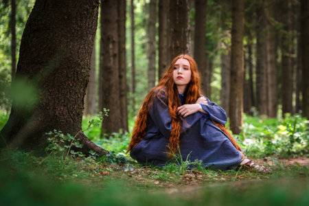 Red haired young woman in a linen blue dress is sitting in the middle of the forest in the rays of the sun. The girl emotions of despair left alone in the forest.の写真素材