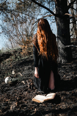 Mystical witch red haired woman in a black long dress in a gloomy sitting forest with book. Gothic...