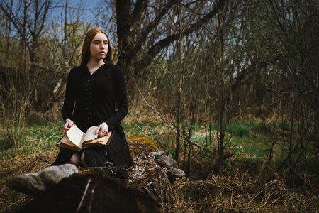 Gothic red haired woman in a black long dress in a gloomy sitting forest with book. Mystical witch...