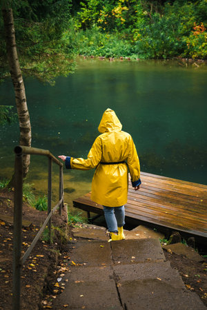Woman in a yellow raincoat and rubber boots descends the stairs to the pier by the lake in a green...
