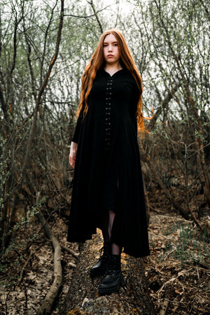 Gothic red haired woman in a black long dress in a gloomy forest. Mystical witch girl.の写真素材