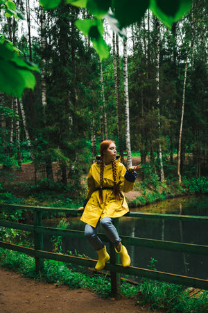 Woman in a yellow raincoat and rubber boots sitting on fence near the lake in a green wet forest during the rain. The concept of travel and tourism.の写真素材