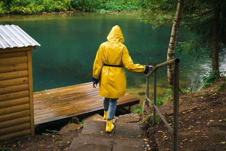 Woman in a yellow raincoat and rubber boots descends the stairs to the pier by the lake in a green wet forest during the rain. The concept of travel and tourism.の写真素材