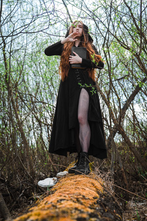 Gothic red haired woman in a black long dress in a gloomy forest. Mystical witch girl.