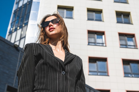 Portrait of a woman in a black jacket and sunglasses posing against the background of an office skyscraper. Businesswoman in the city.の写真素材