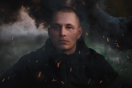 Portrait of a serious sad man in the smoke of a black smoke bomb close-up in a field.