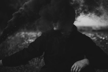 Portrait of a serious sad man in the smoke of a black smoke bomb close-up in a field. Black and...