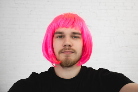 Selfie of a bearded man in a pink wig in close up on a white wall background.の写真素材