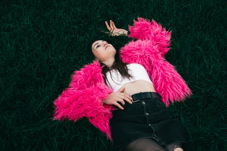 Extravagant woman in a pink fur coat and torn tights lies on a lawn of green grass.の写真素材