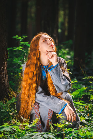 Red haired young woman in a linen blue dress is sitting in the middle of the forest in the rays of...