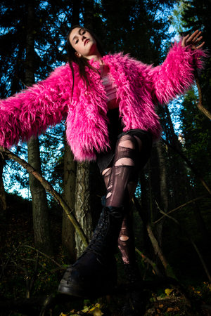An eccentric young woman poses in a pink fur coat and torn tights in a dark green forest.の写真素材