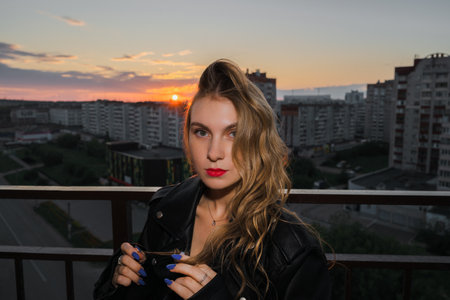 Woman in black casual clothes stands on a balcony in the light of the setting sun against the background of a small city street.の写真素材