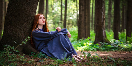 Red haired young woman in a linen blue dress is sitting under a tree of the forest in the rays of the sun. The girl emotions of despair left alone in the forest.の写真素材