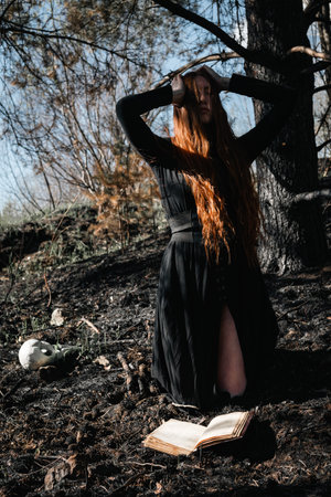 Mystical witch red haired woman in a black long dress in a gloomy sitting forest with book. Gothic girl on a scorched ground.の写真素材