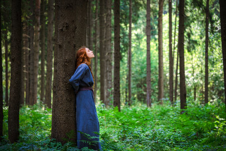 Red haired young woman in a linen blue dress is escape in the middle of the forest in the rays of...