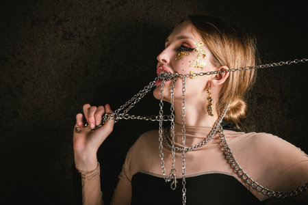 Portrait of a drunken woman in chains with a gold make-up on a dark background. Tipsy girl is trying...