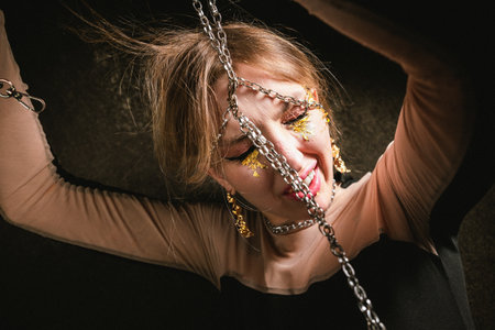 Portrait of a drunken woman in chains with a gold make-up on a dark background. Tipsy girl is trying...