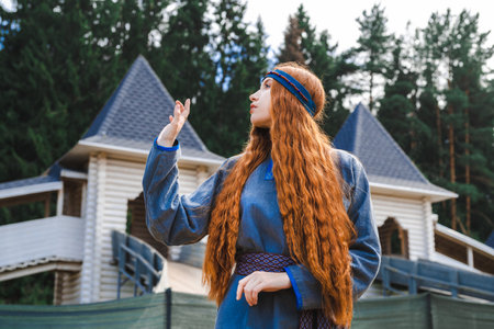 Portrait of a red haired woman in a blue medieval linen dress thinking on the background of a wooden castle.の写真素材