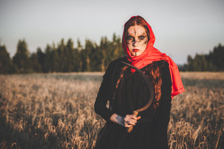 Angry witch in a red headscarf and a black robe stands in a field of wheat with a sickle. Concept of the curse and the harvest.の写真素材