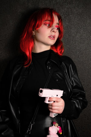 Young woman with red hair on a black background. A girl in black leather clothes holds a pink pistol in her hand. The concept of self-defense or self-protection.の写真素材