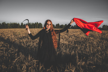 Angry witch in a red headscarf and a black robe standing in a field of wheat with a sickle. Concept...