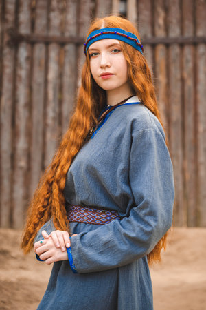Portrait of a red haired woman in a blue medieval linen dress serious on the background of a wooden castle gate.の写真素材