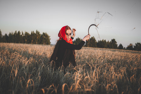 Angry witch in a red headscarf and a black robe standing in a field of wheat with a sickle. Concept...