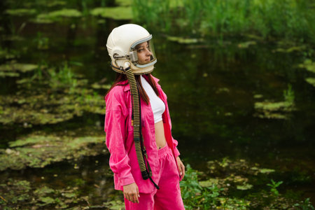 Woman in an astronaut space helmet and a pink suit in a wild green forest by a pond.
