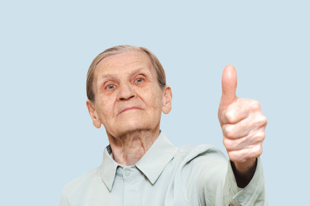 An elderly woman gives a thumbs-up with a calm, confident expression, wearing a light-colored shirt...