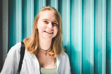 A young woman smiling in front of a turquoise wall, wearing a white shirt and a necklace.