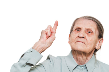 An elderly woman with a serious expression points her finger upward, wearing a light-colored shirt on a white background.の写真素材