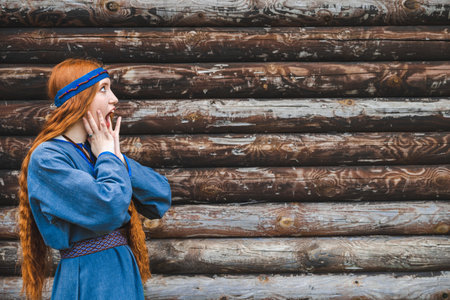 Red haired woman with freckles in a blue linen medieval dress against the background of a wooden fortress palisade.の写真素材