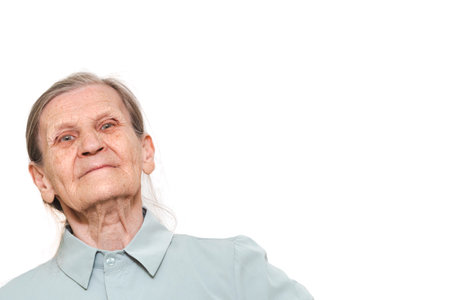 An elderly woman with a gentle smile and calm expression, wearing a light shirt against a plain...