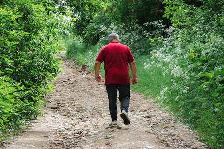 A man in a red shirt walks alone down a rugged path, surrounded by lush greenery.の写真素材